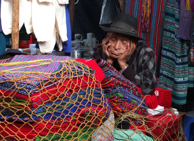 Hombre junto a telas típicas de Guatemala, aparentemente con sueño o cansado, con los ojos cerrados. Vistiendo trajes tipicos de guatemala y un sombrero negro